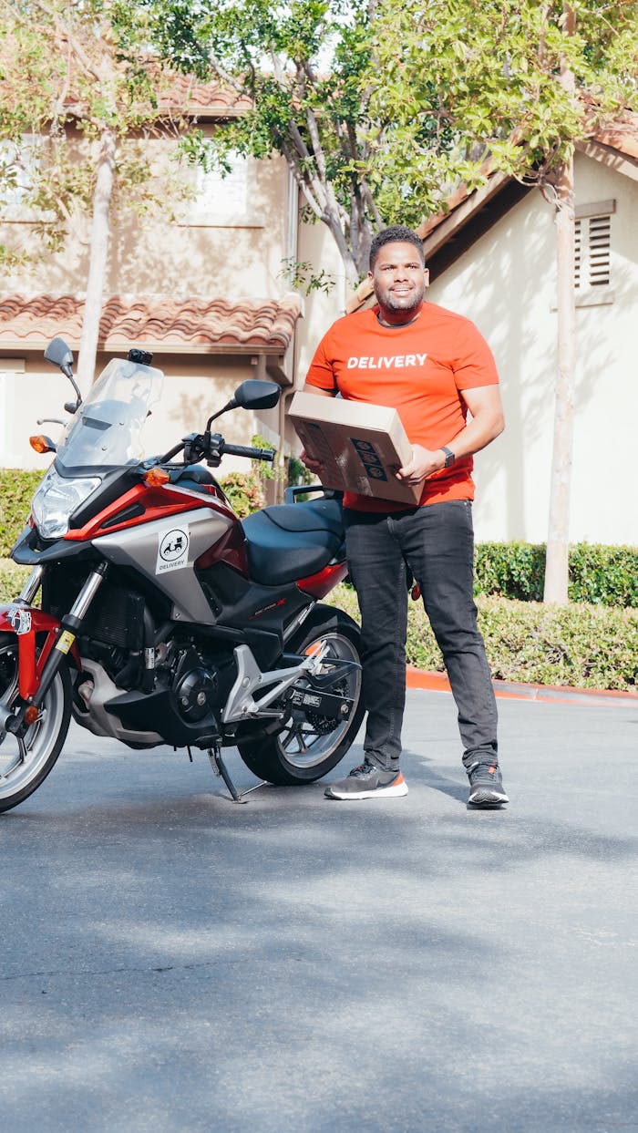 gallery-2 A delivery man in a red uniform holding a package beside a motorcycle in an outdoor setting.